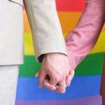 two men holding hands against a Pride flag
